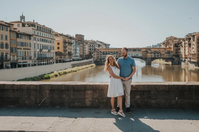 The couple stand close together by the Arno with Ponte Vecchio framed behind them during their Florence couple photoshoot.