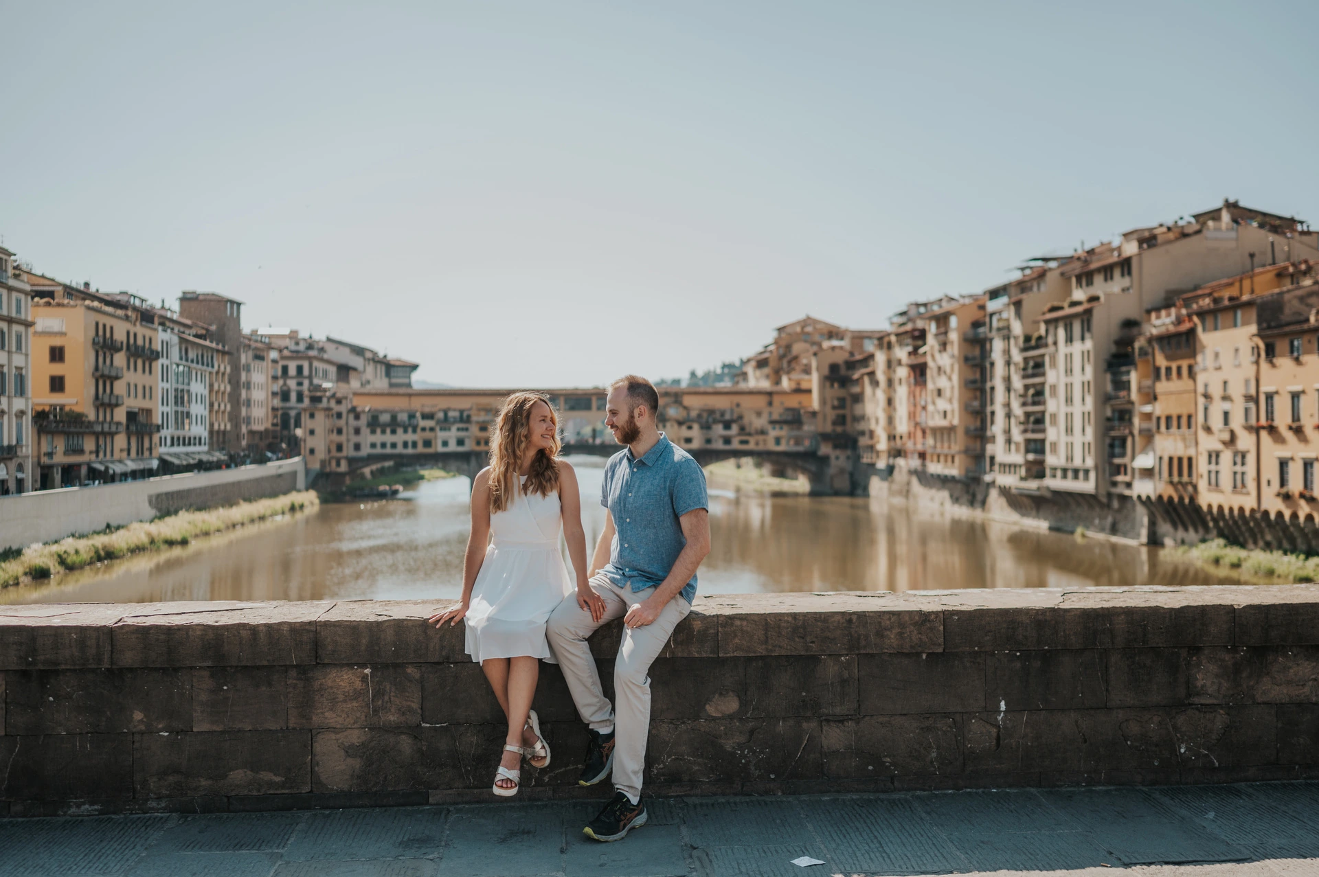 The couple lean together by the stone river parapet, with Ponte Vecchio behind them, during their Florence photoshoot.