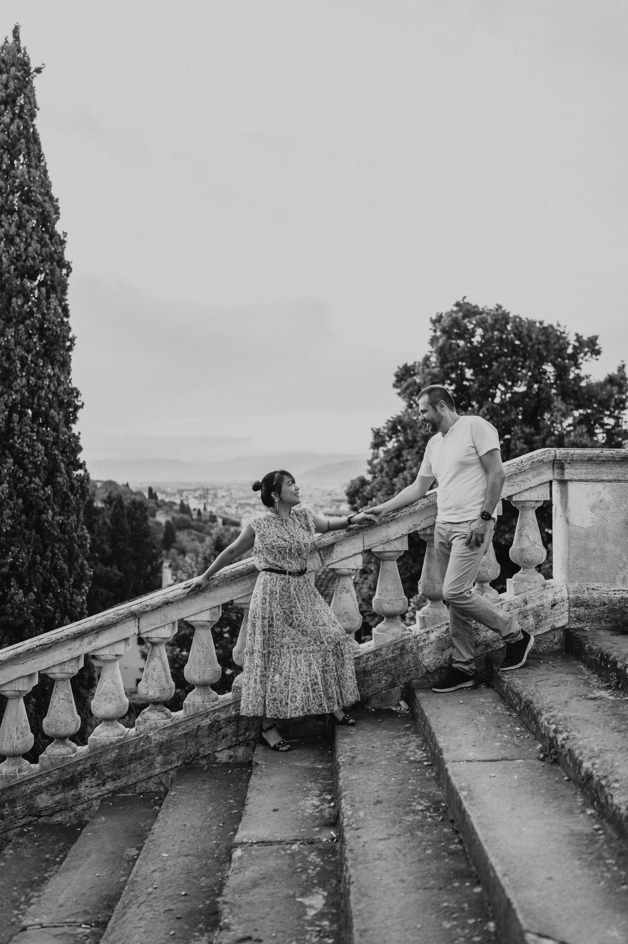 Black-and-white full-length portrait of the couple holding hands on the stone steps during their Florence photoshoot, with trees above the balustrade.