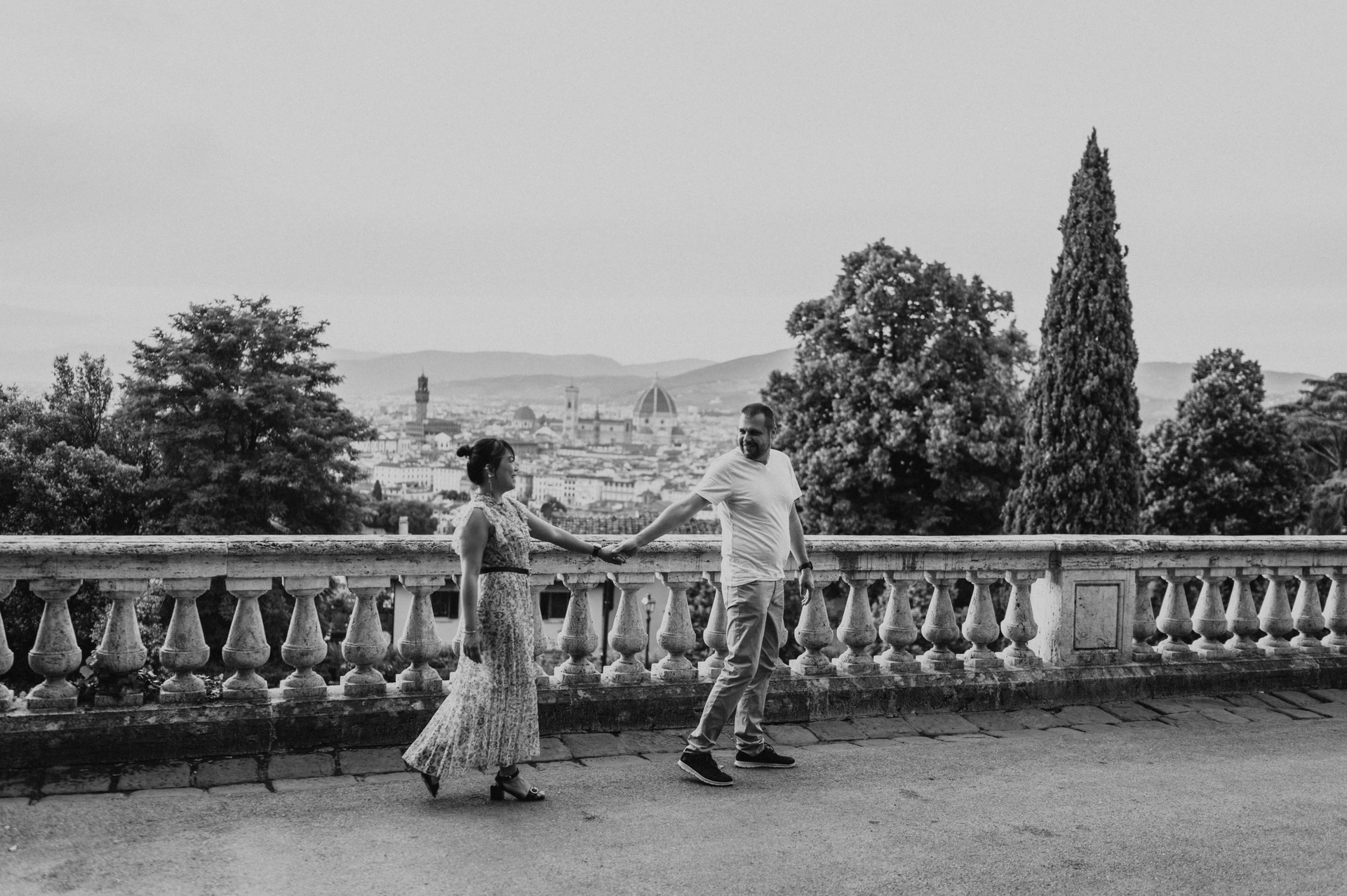 Black-and-white Florence photoshoot of the couple walking hand in hand along the balustrade, with trees and the city panorama behind them.