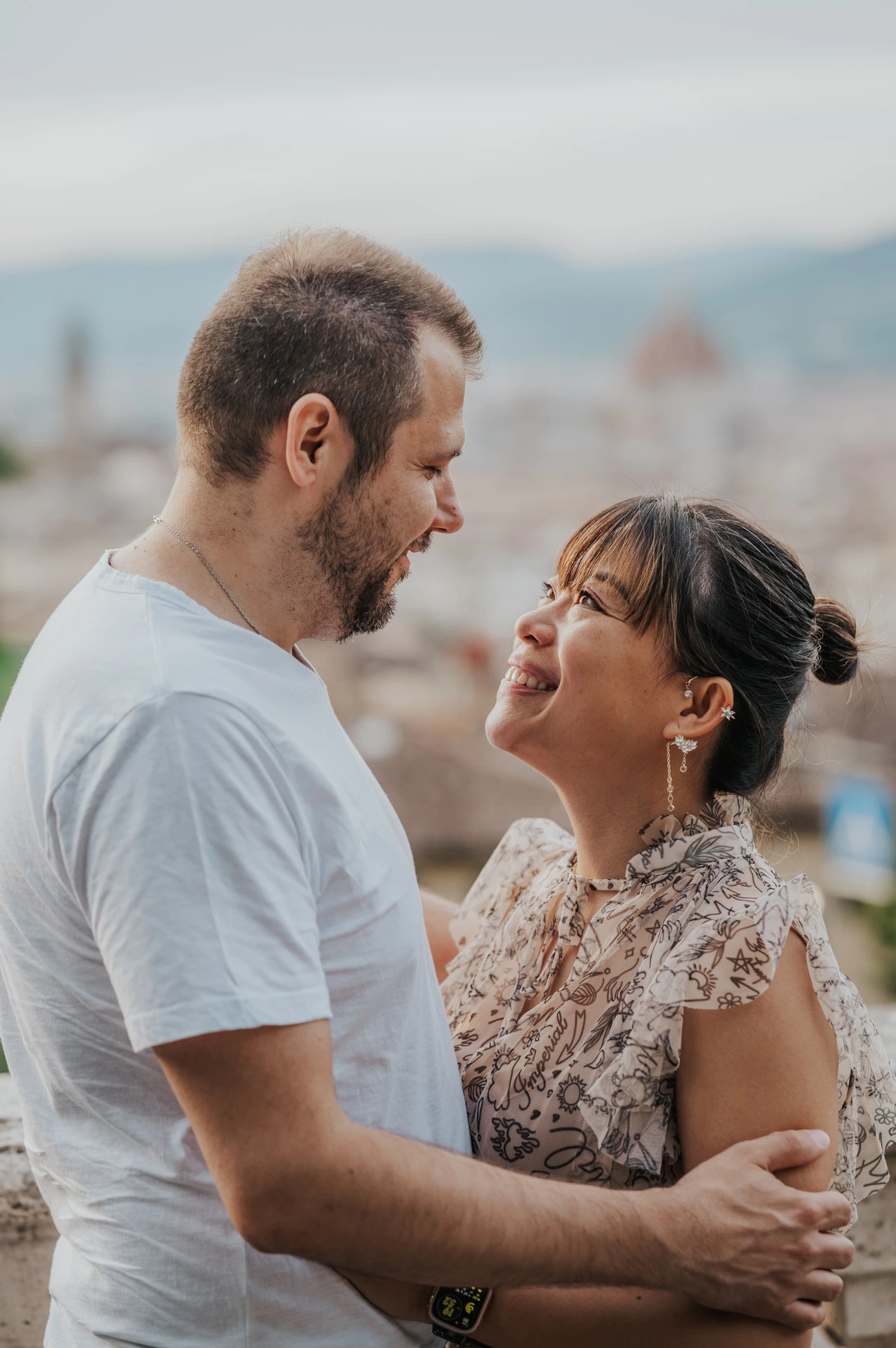 The couple smile at each other in a close Florence photoshoot portrait, with pale rooftops and the cathedral dome blurred behind them.