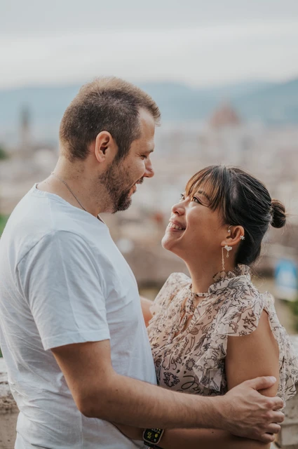 The couple smile at each other in a close Florence photoshoot portrait, with pale rooftops and the cathedral dome blurred behind them.