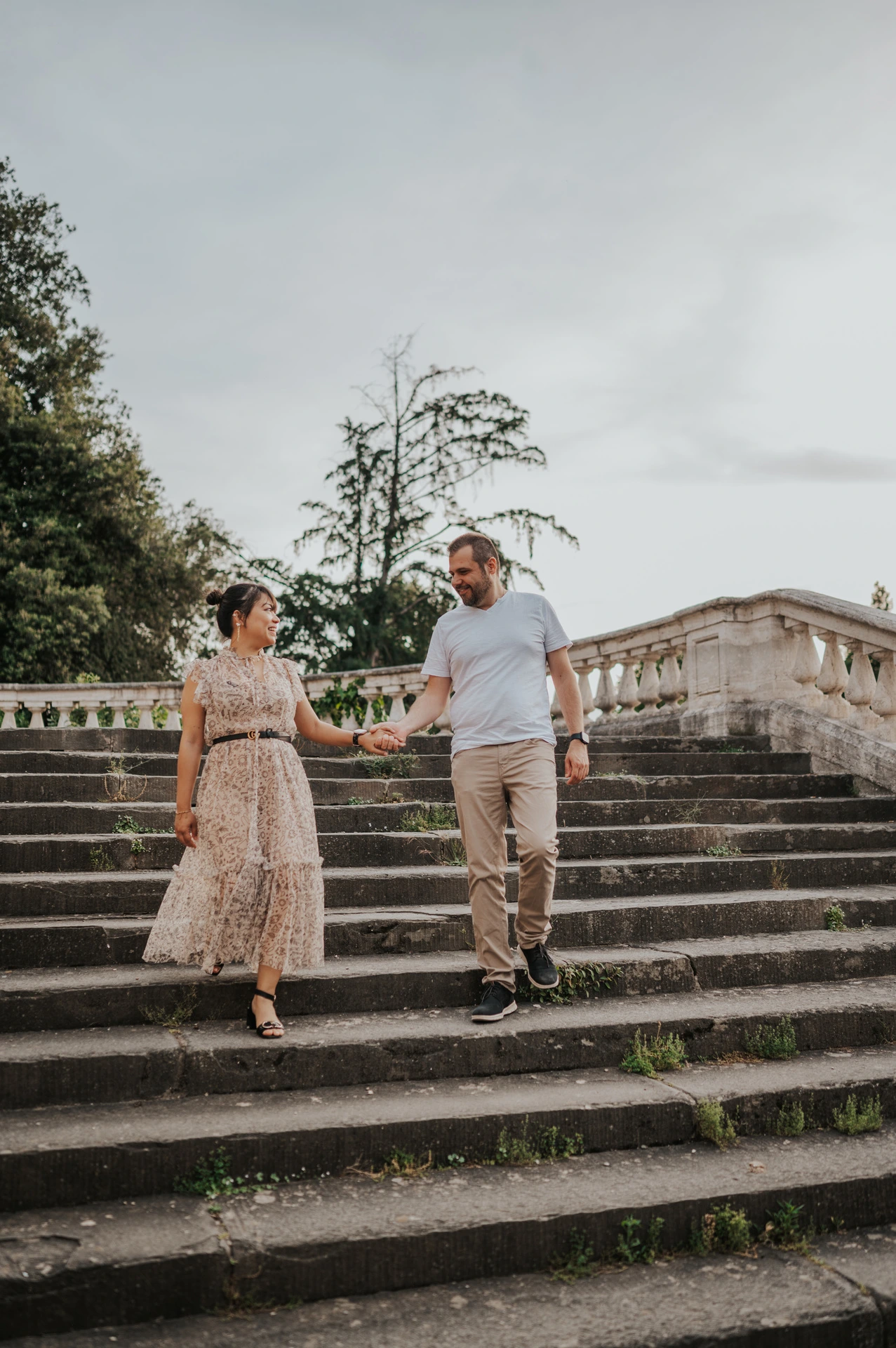 The couple walk down the broad stone steps holding hands during their Florence photoshoot, with the balustrade rising behind them.