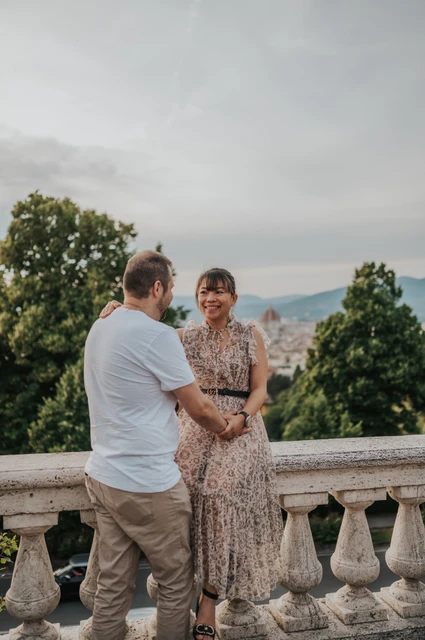 Florence photoshoot holding hands by the balustrade The woman smiles towards the man as they hold hands beside the balustrade in their Florence photoshoot, with trees and rooftops behind them.