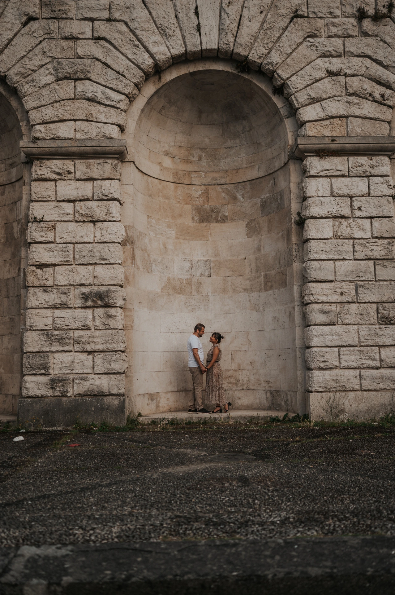 The couple kiss beneath the tall arched stone recess during their Florence photoshoot, with the stonework and gravel foreground in view.