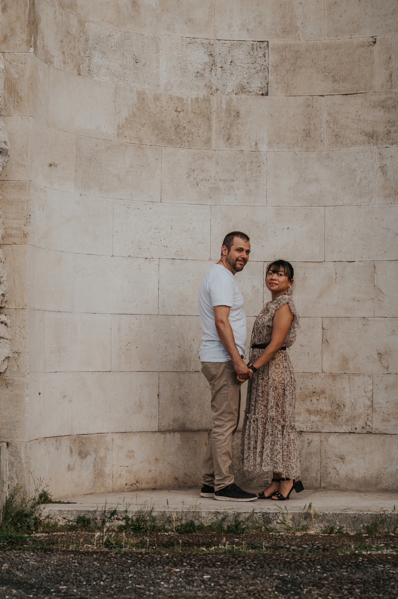 The couple stand close together and smile towards the camera against the pale stone wall of an arched recess during their Florence photoshoot.
