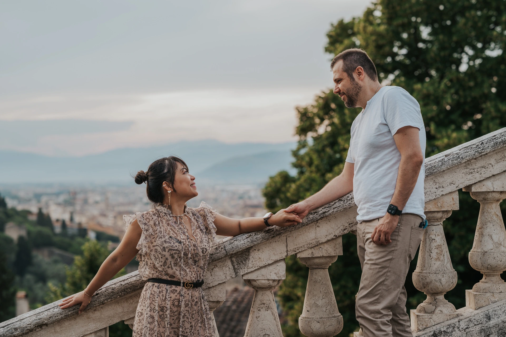 The couple hold hands beside the sloping stone stair rail during their Florence photoshoot, with Florence rooftops visible between the trees.