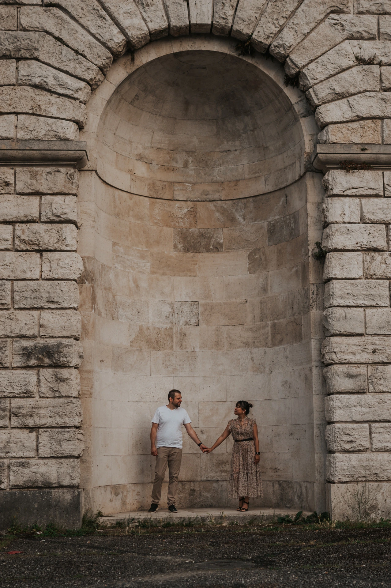 The couple hold hands at the base of a tall arched stone recess in their Florence photoshoot, with the full stonework filling the portrait.