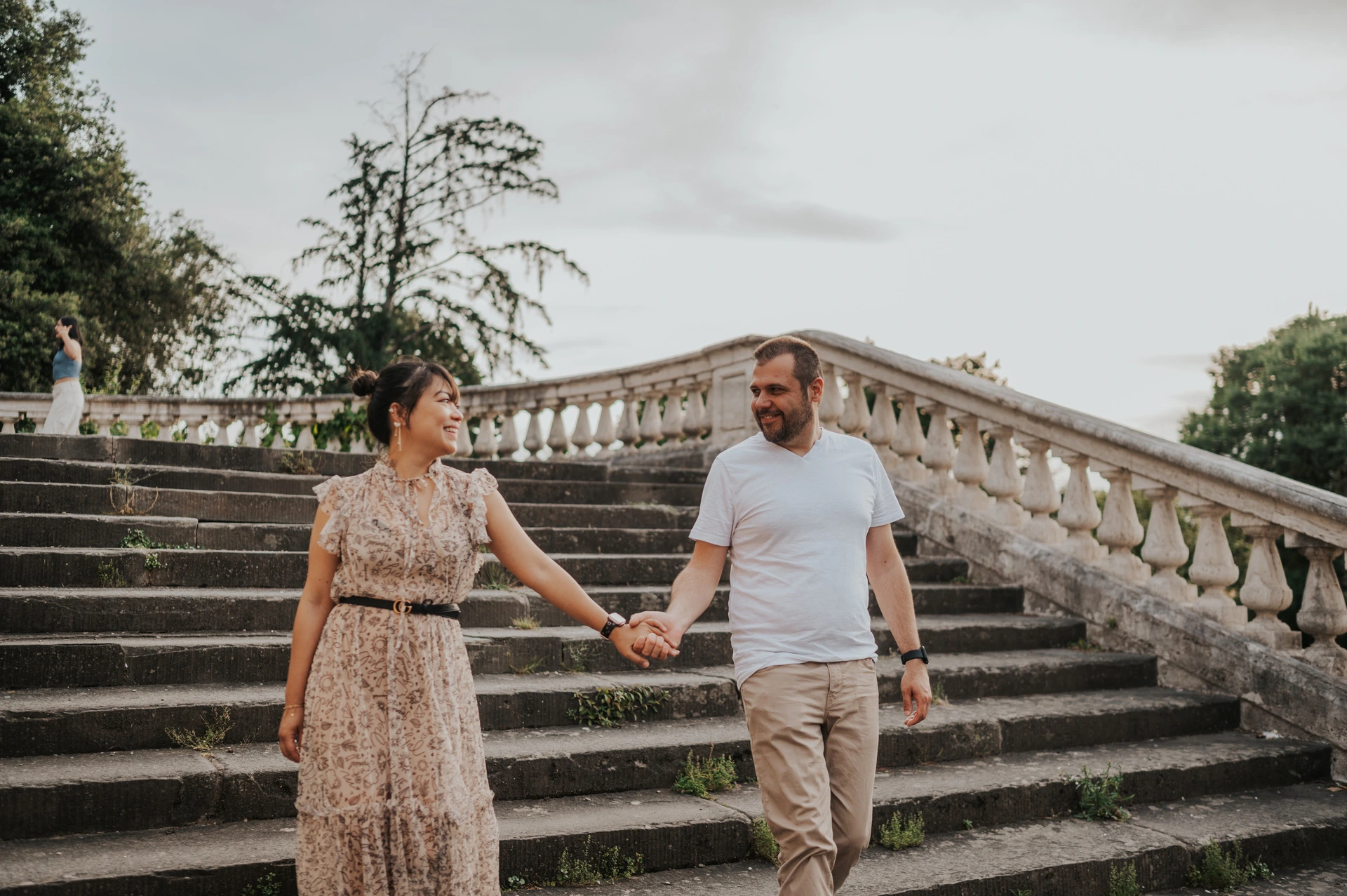 The couple hold hands as they walk down the stone staircase in a landscape Florence photoshoot portrait, with trees behind the balustrade.
