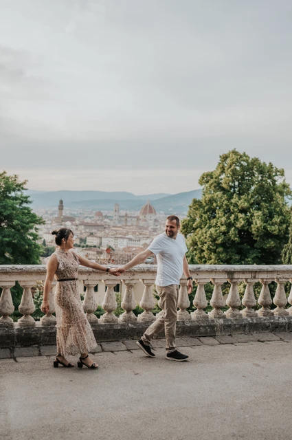 The couple walk hand in hand beside the stone balustrade during their Florence photoshoot, with trees and the city skyline behind them.