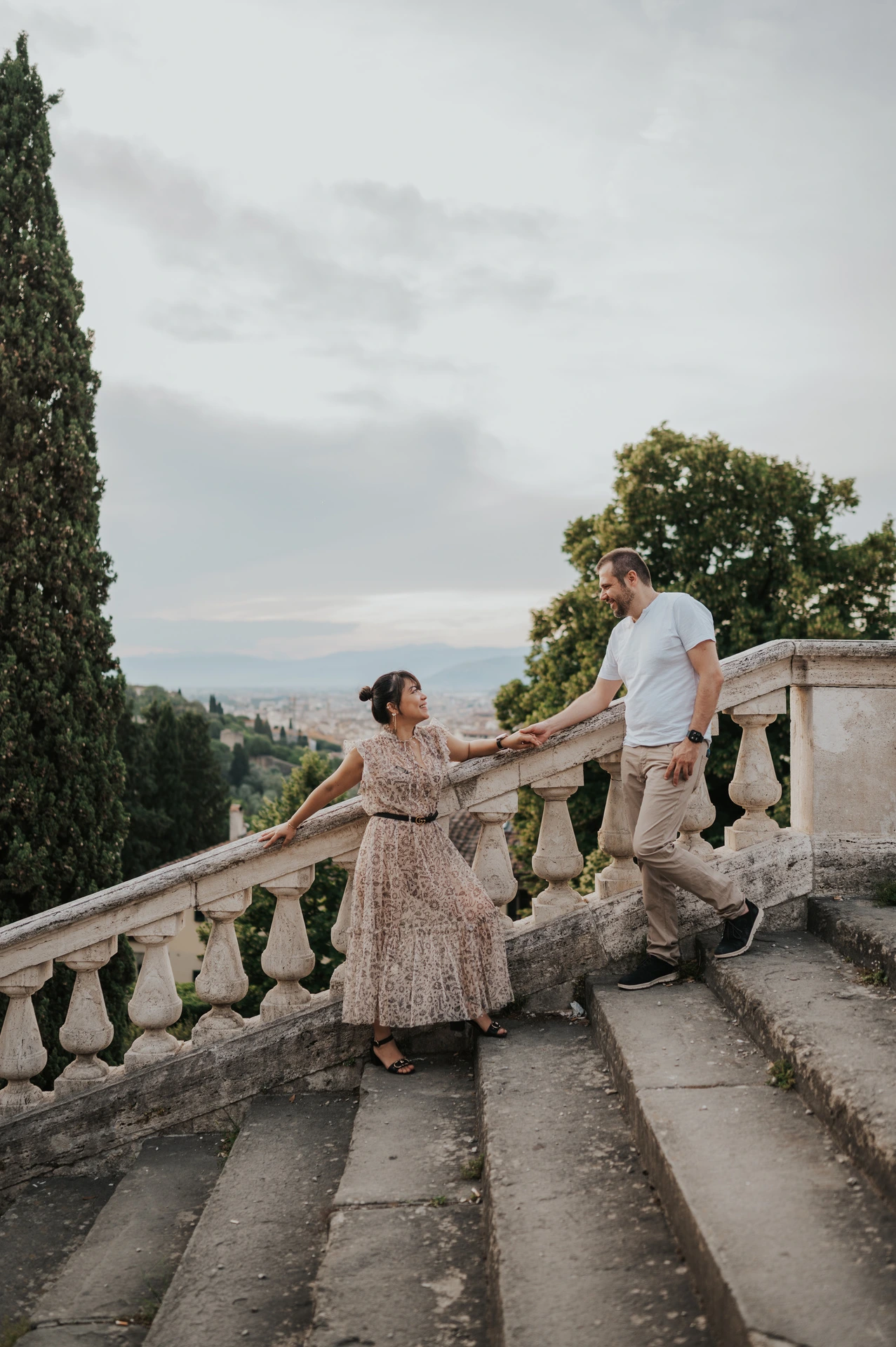 The couple stand at different levels on the stone stairs for their Florence photoshoot, with a tall cypress and the balustrade beside them.