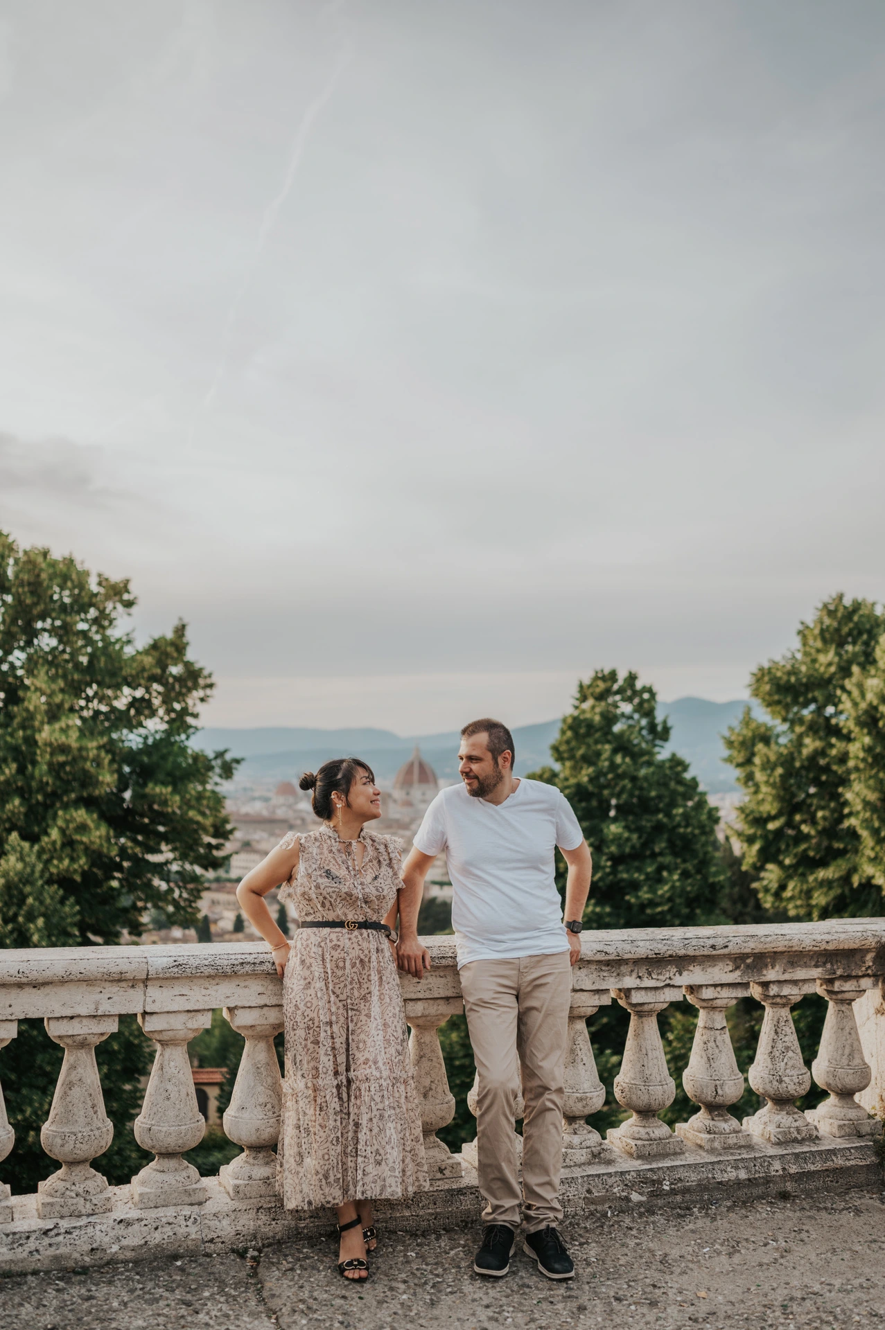 The couple lean beside the stone balustrade in a full-length Florence photoshoot portrait, framed by trees and the city below.