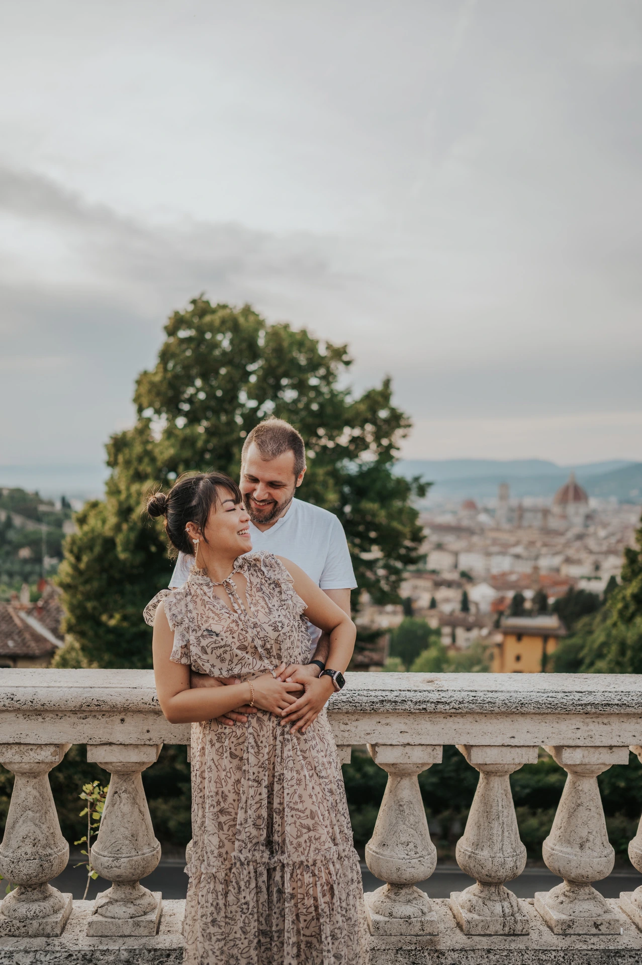 The couple stand in an embrace by the balustrade during their Florence photoshoot, with their full outfits, trees and city rooftops in view.