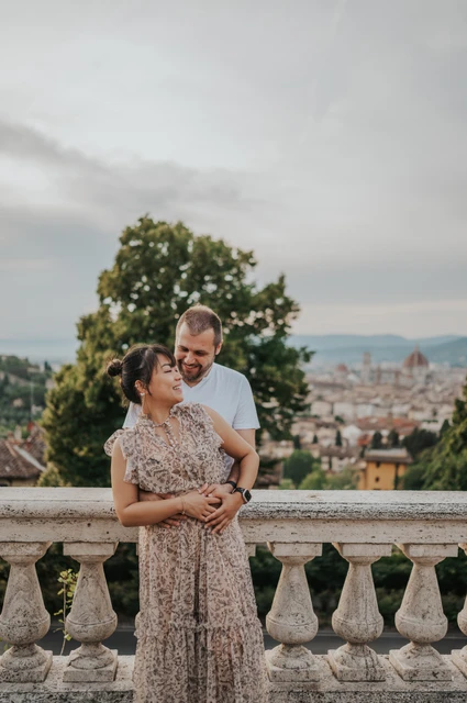 Florence photoshoot embrace at the stone balustrade The couple stand in an embrace by the balustrade during their Florence photoshoot, with their full outfits, trees and city rooftops in view.