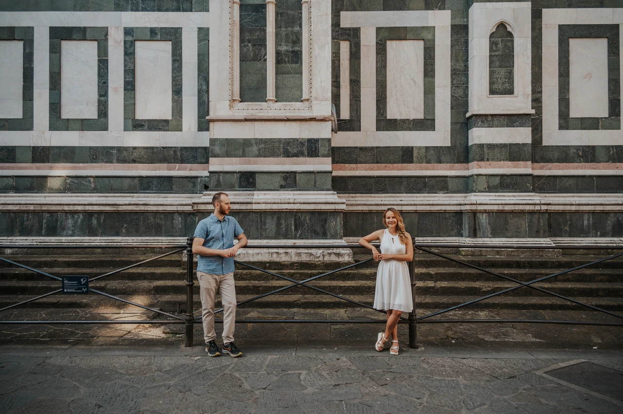 A wide striped-marble view from a Florence couple photoshoot The couple stand apart by a railing beneath the cathedral’s striped marble wall during their Florence couple photoshoot.