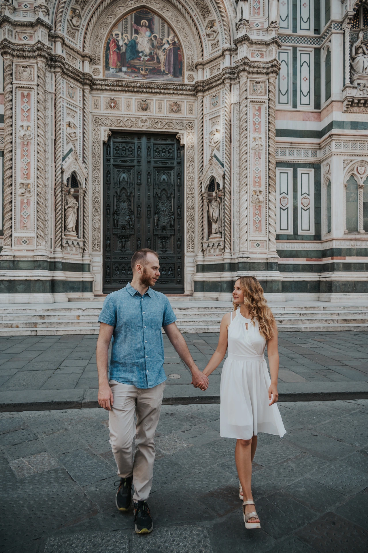 The couple walk together in front of the Duomo’s marble entrance, wearing a blue shirt and white dress, during their Florence photoshoot.