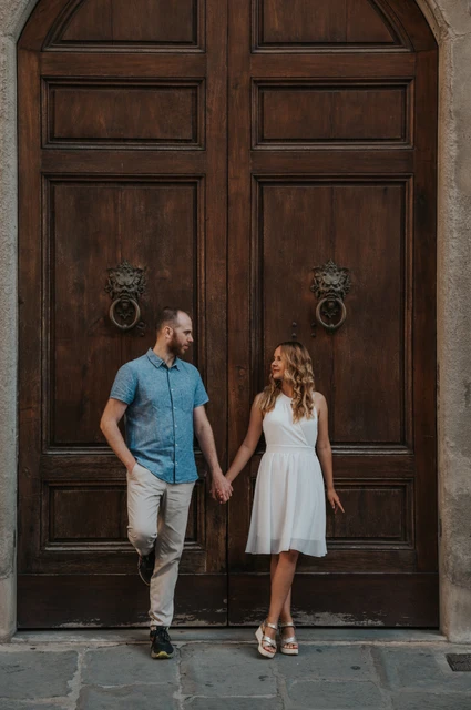 A walking portrait by a wooden door during a Florence photoshoot The couple walk hand in hand in front of a dark wooden door during their Florence couple photoshoot.