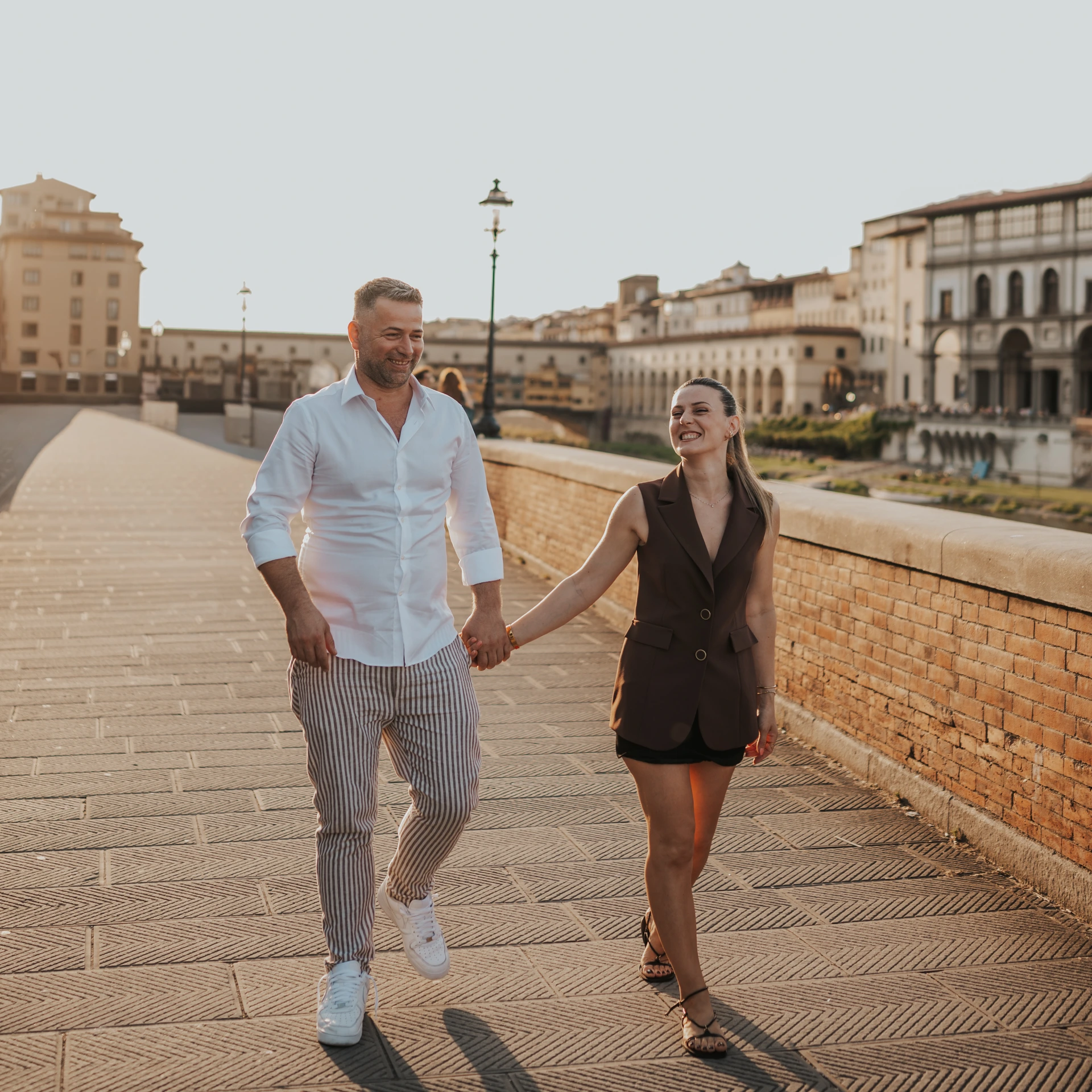 Una coppia sorridente cammina mano nella mano lungo il parapetto in mattoni di un ponte a Firenze.