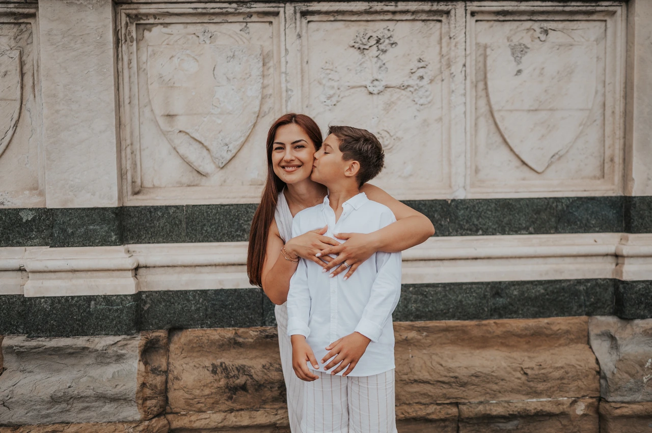 A kiss on the cheek during a Florence family photoshoot The boy kisses the woman on the cheek as they stand by the marble wall during their Florence family photoshoot.