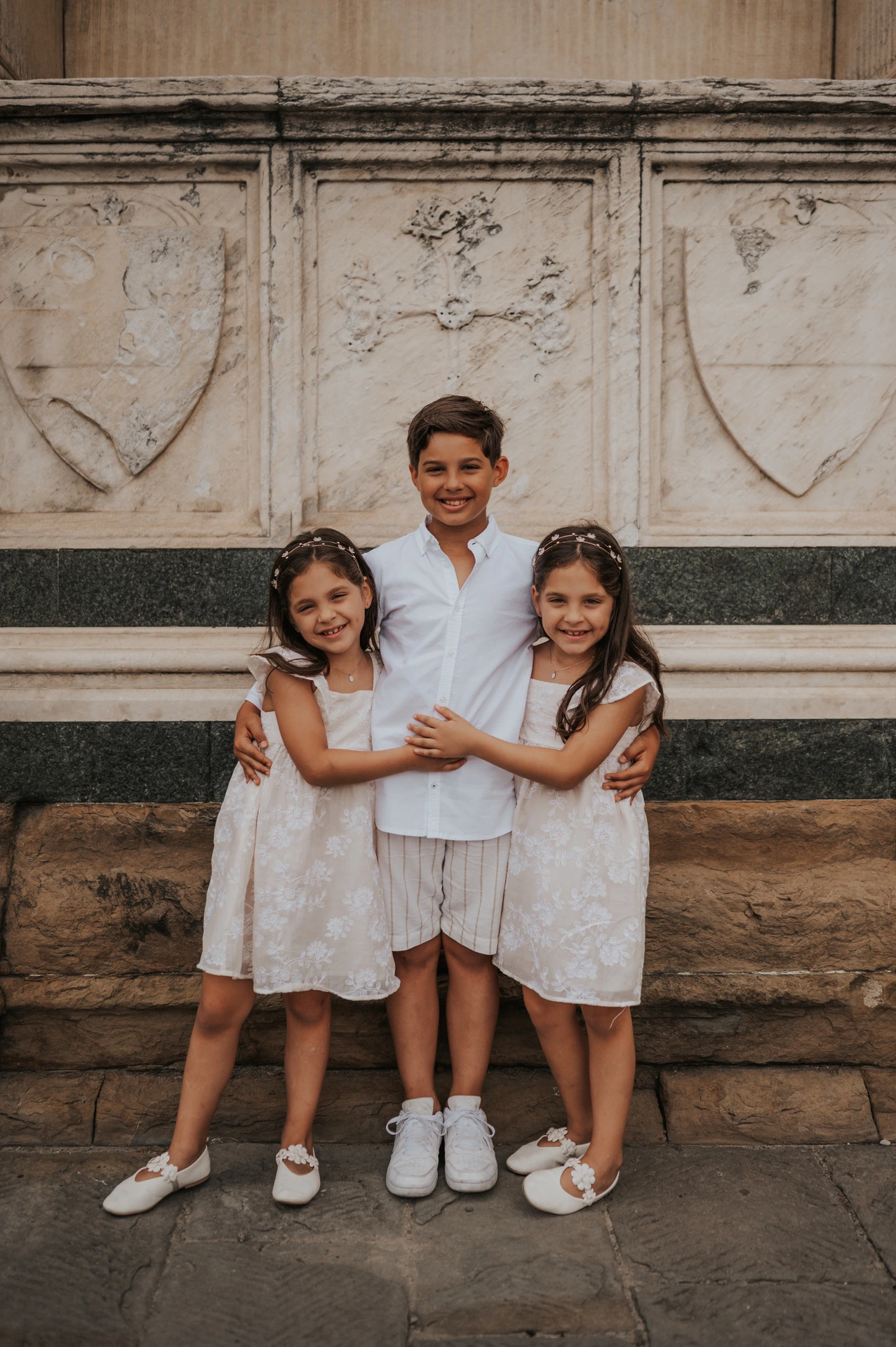The three children hug in front of a carved marble wall and stone ledge during their Florence family photoshoot.