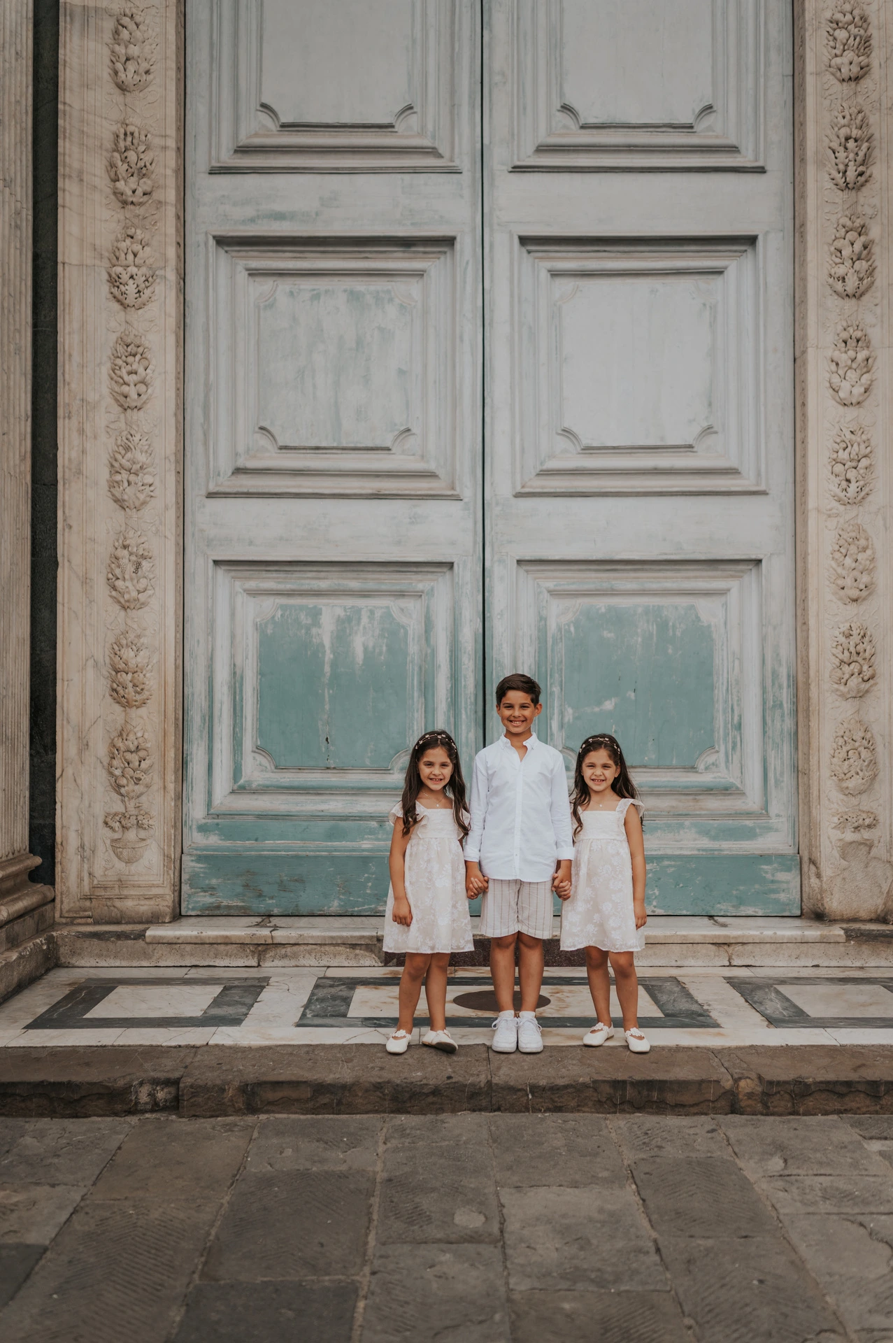 Three children stand holding hands below a tall green door during their Florence family photoshoot.