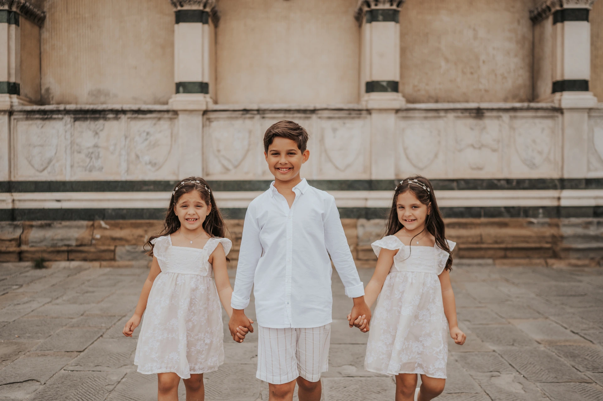 Florence family photoshoot with the children walking together The three children walk forward holding hands, wearing white, during their Florence family photoshoot beside the marble wall.