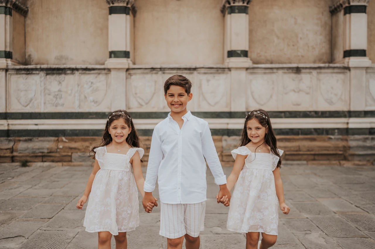 Florence family photoshoot with the children walking together The three children walk forward holding hands, wearing white, during their Florence family photoshoot beside the marble wall.