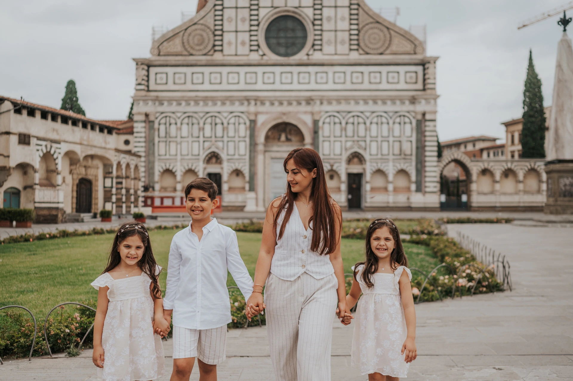 The family hold hands beside flowerbeds with Santa Maria Novella’s patterned facade behind them during their Florence photoshoot.