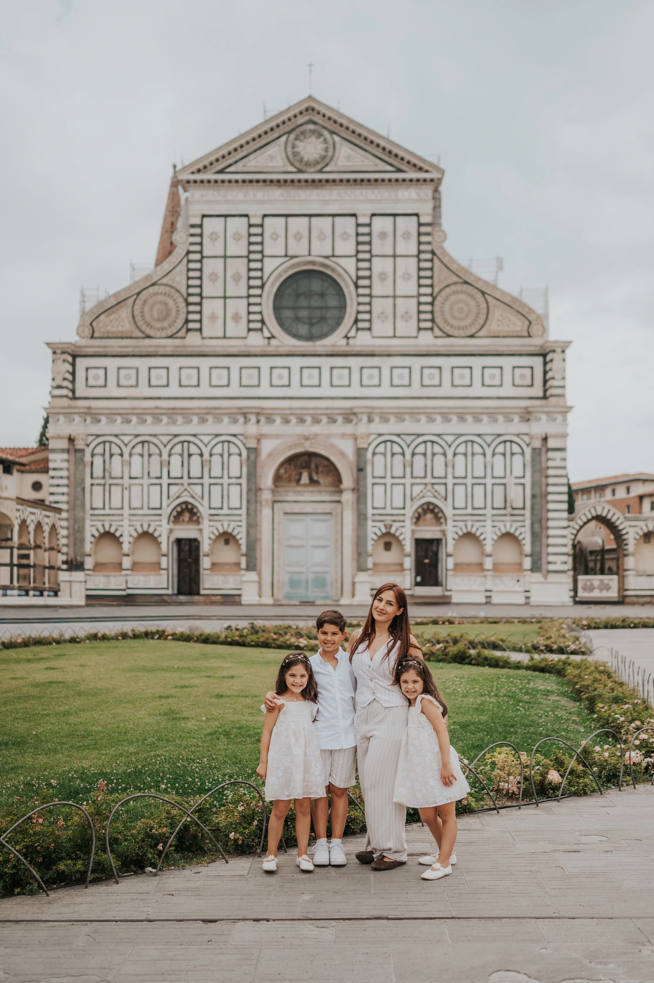 The family walk together on the square beside the flowerbeds, with Santa Maria Novella behind them, during their Florence family photoshoot.