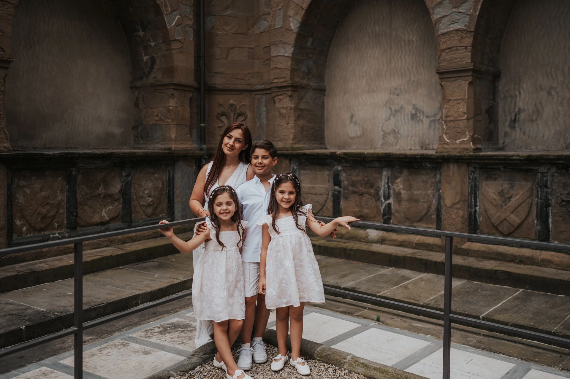 The family stand together by a metal railing and weathered stone arches during their Florence photoshoot.