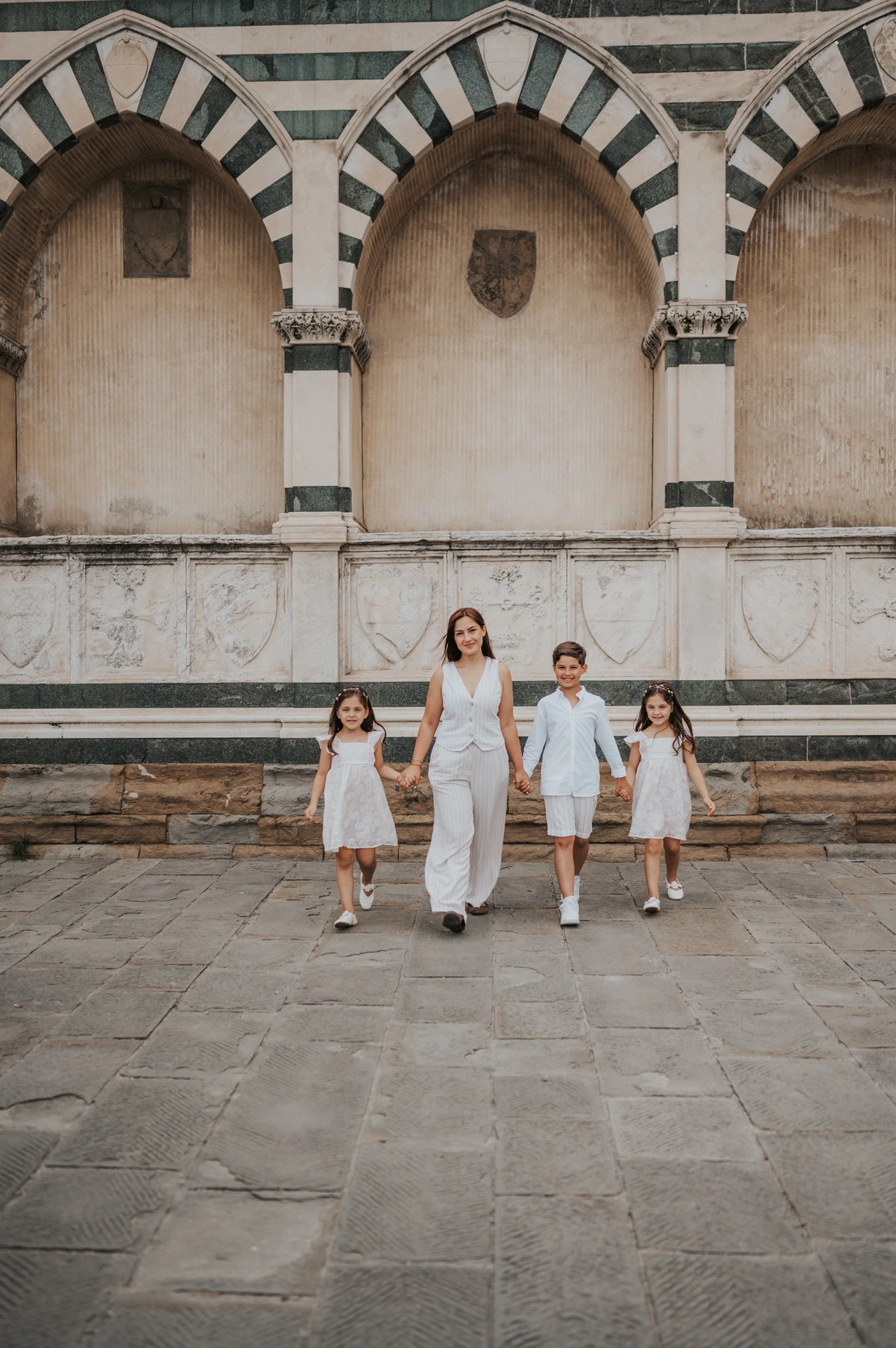 The family of four walk across the stone paving beneath striped arches during their Florence family photoshoot.