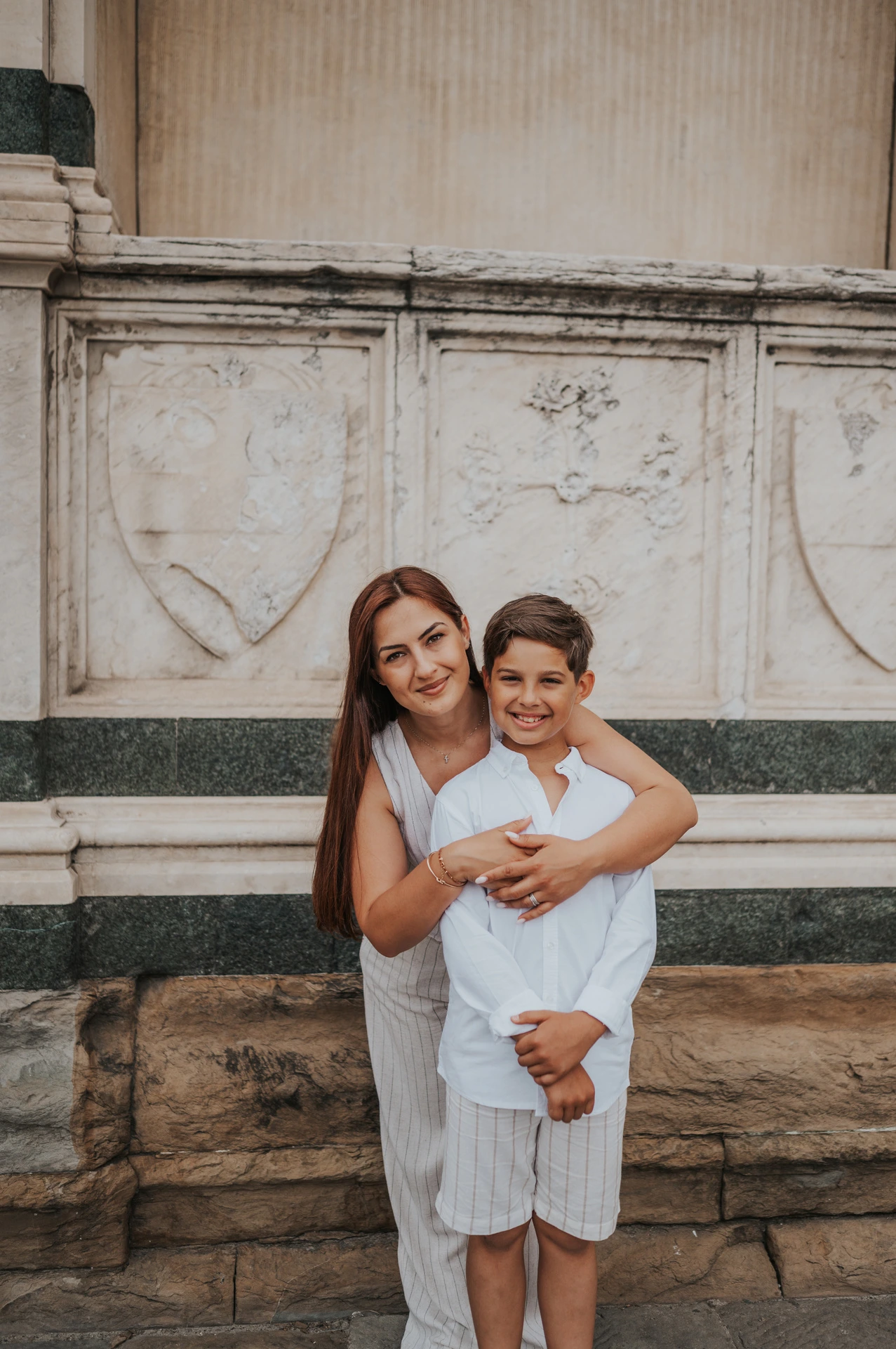 A woman and boy smile together beside the marble wall during their Florence family photoshoot.