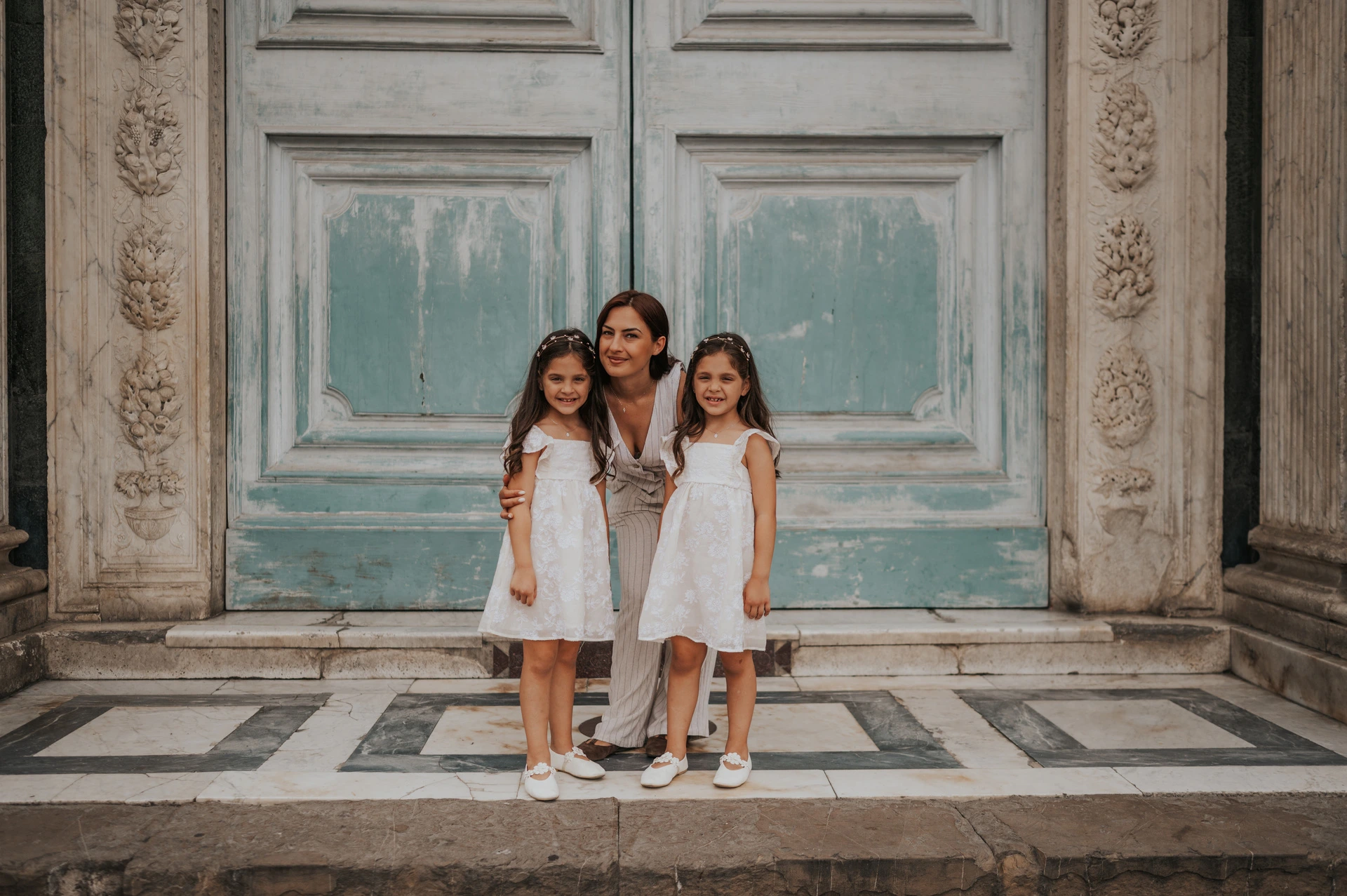 A woman crouches with two girls in white dresses in front of the marble doorway during their Florence family photoshoot.