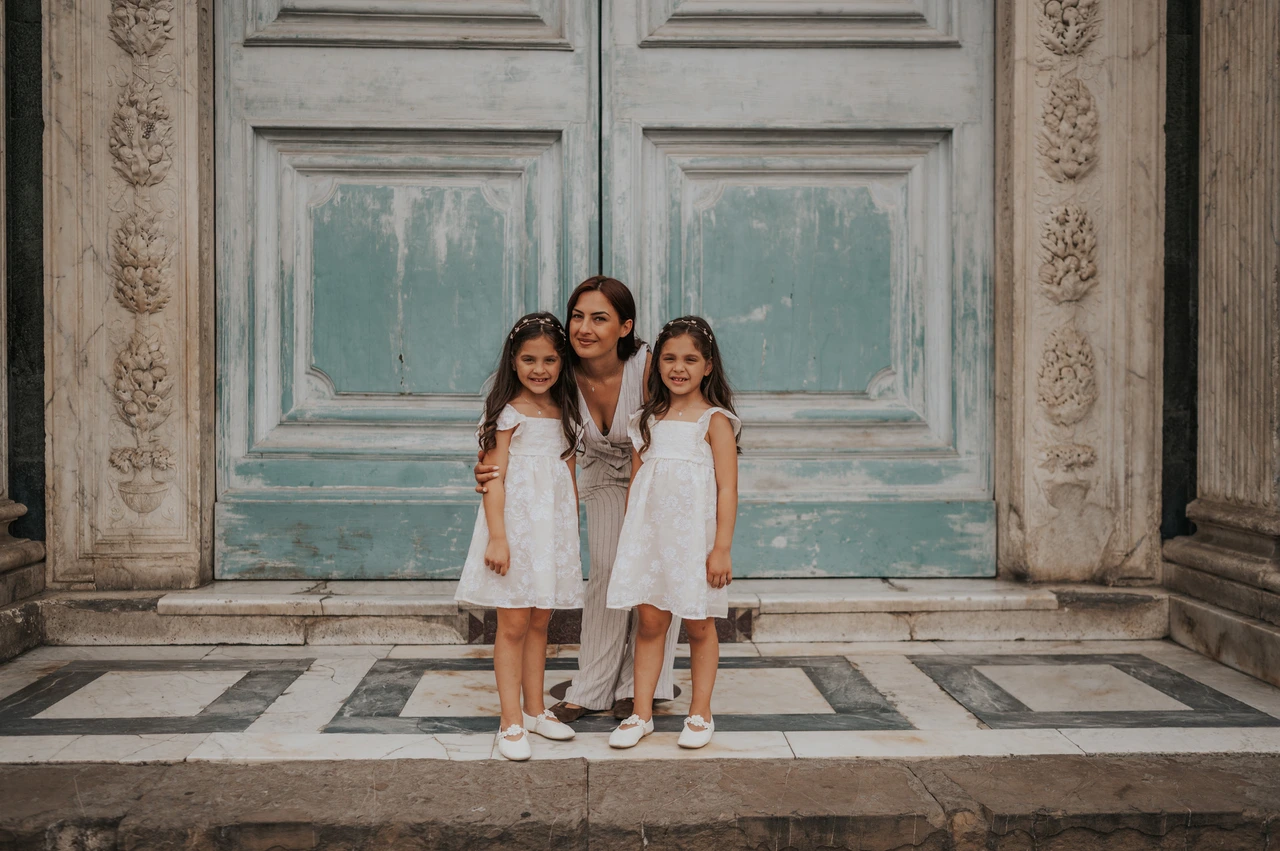 Florence family photoshoot with three smiling faces at the doorway A woman crouches with two girls in white dresses in front of the marble doorway during their Florence family photoshoot.