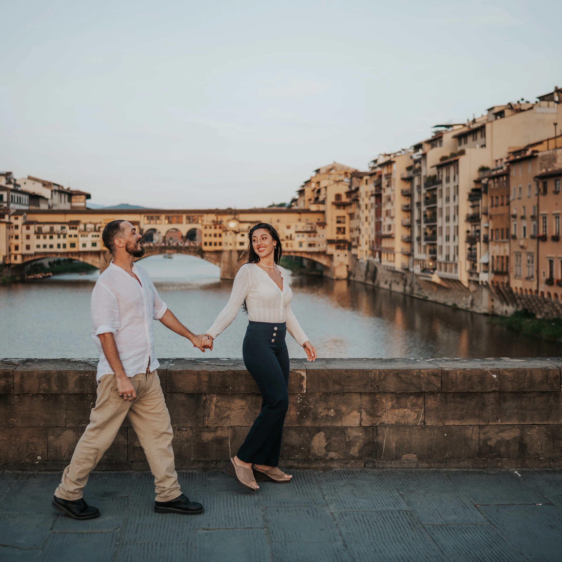 Una coppia si tiene per mano lungo l’Arno, con Ponte Vecchio e gli edifici sul fiume alle spalle.