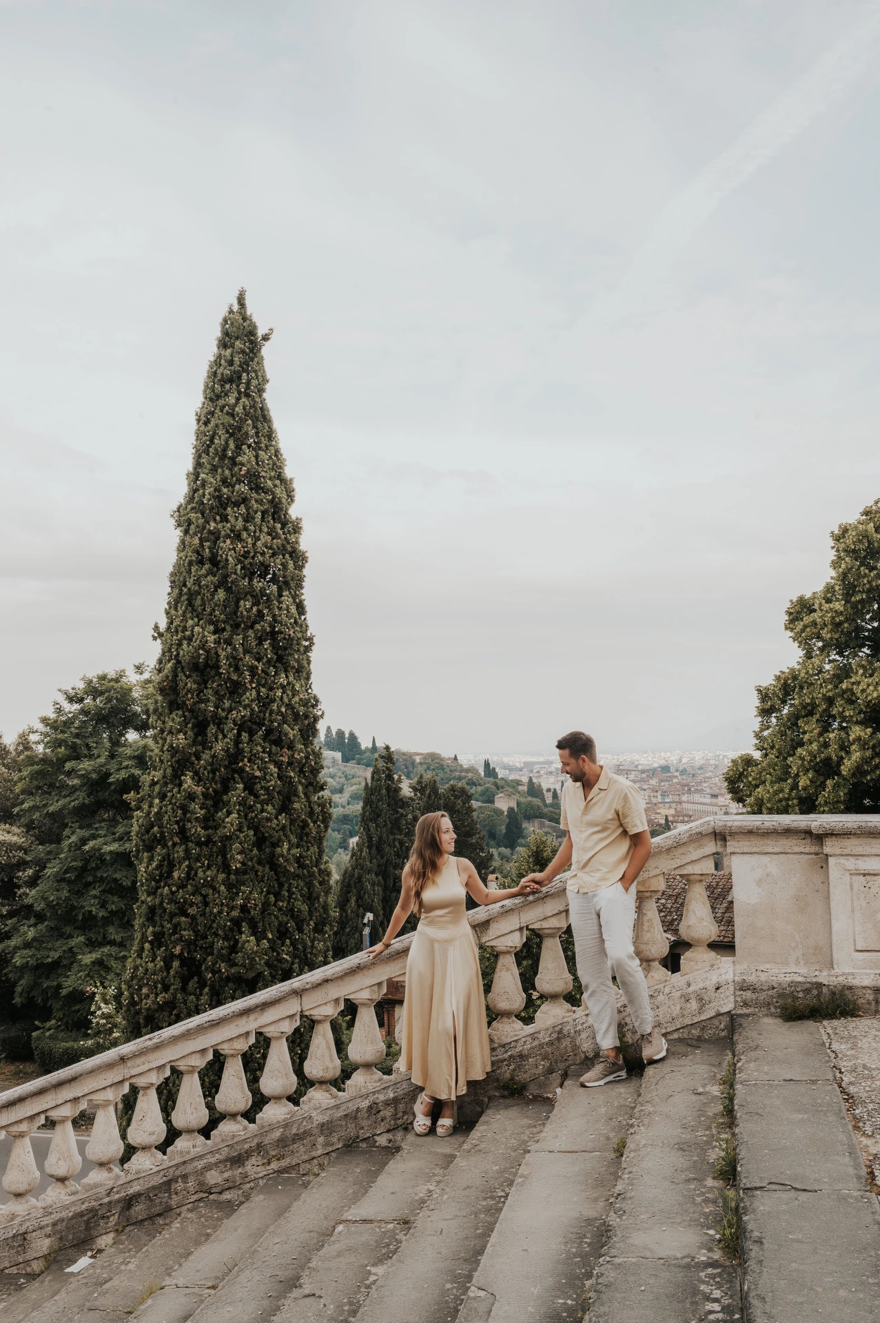The couple hold hands by broad stone steps and a tall cypress tree during their Florence proposal photoshoot.