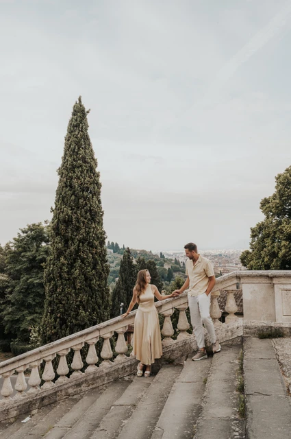 The couple hold hands by broad stone steps and a tall cypress tree during their Florence proposal photoshoot.