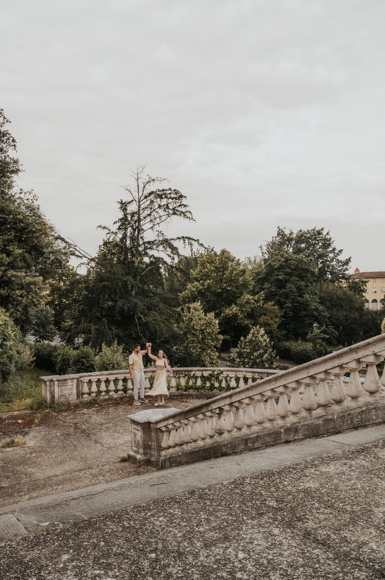 The couple stand together beyond a stone stairway balustrade, surrounded by greenery, during their Florence proposal photoshoot.