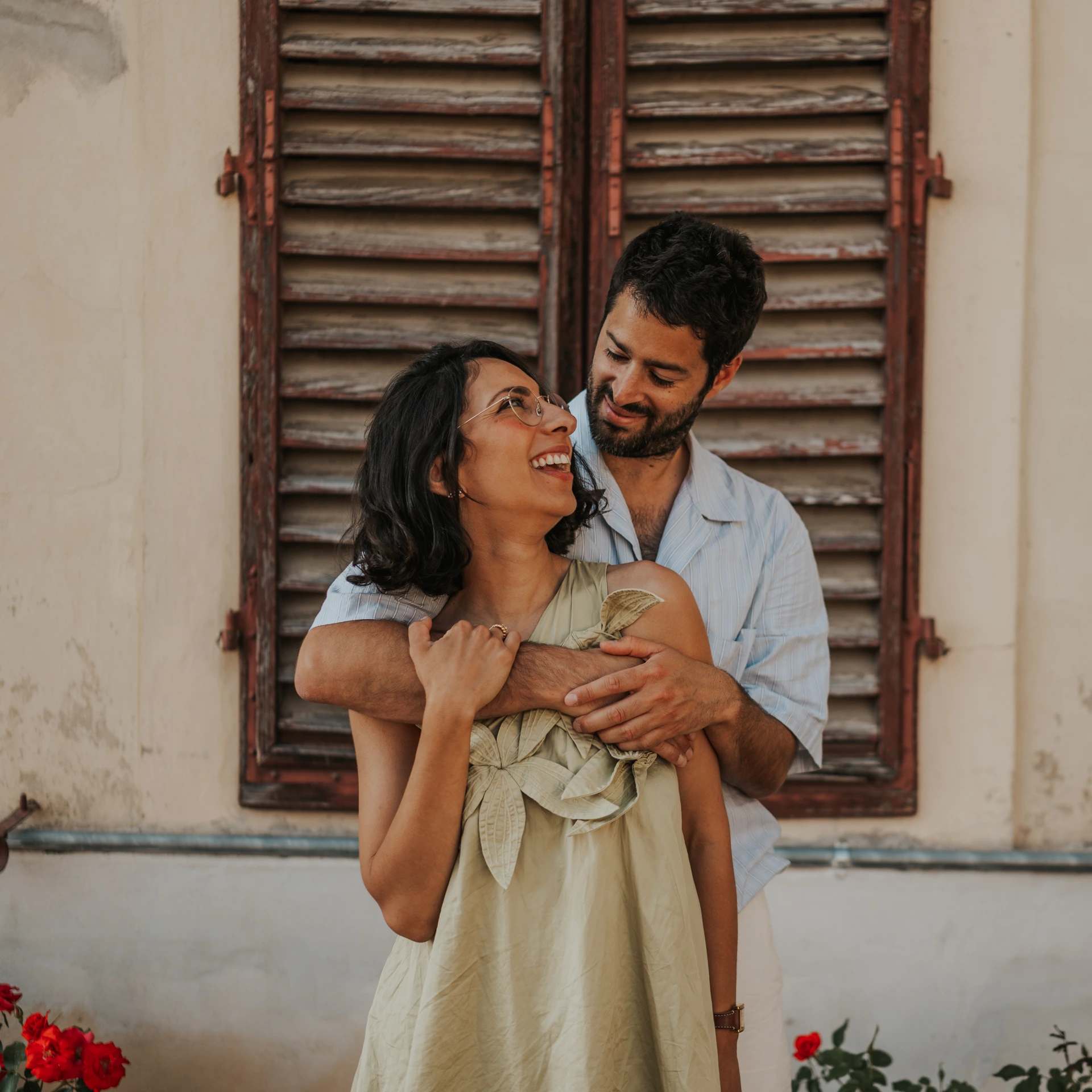 Una coppia sorridente si abbraccia davanti a persiane in legno vissuto e a una parete chiara.