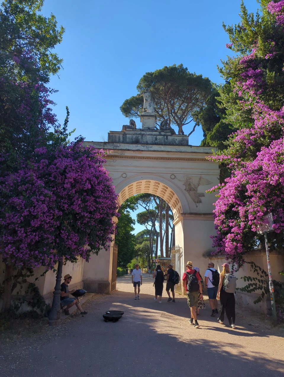 Rome Summer Photoshoot Outfits - Garden Arch Visitors in light summer clothes and sun hats walk beneath a flower-framed arch on a bright day in Rome.