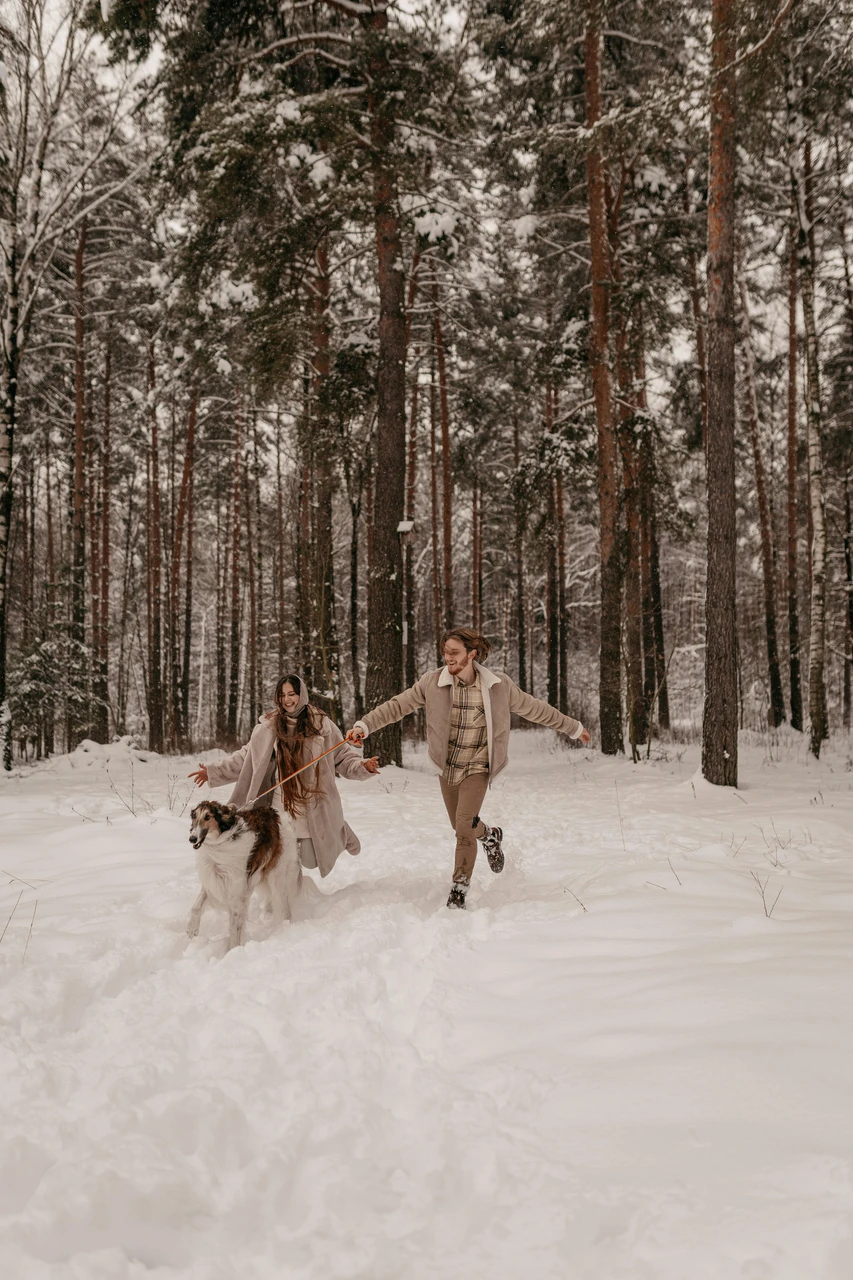 Outfits that stand out from forest tones in engagement photos Couple and dog running through snowy forest in coordinated outfits