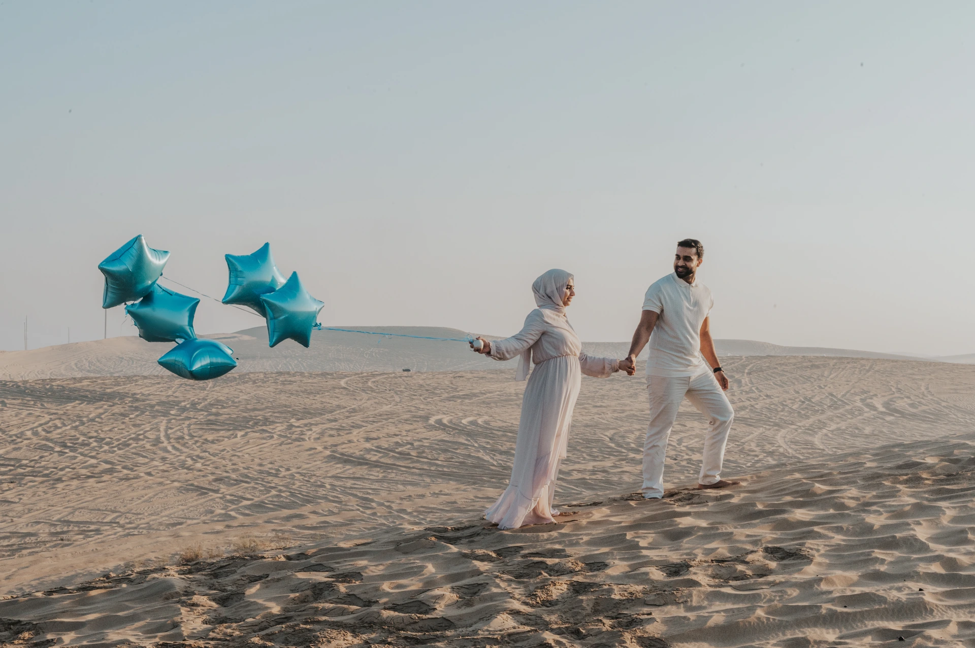 A couple walks hand in hand on Dubai desert dunes while carrying blue star balloons during a gender reveal photoshoot.