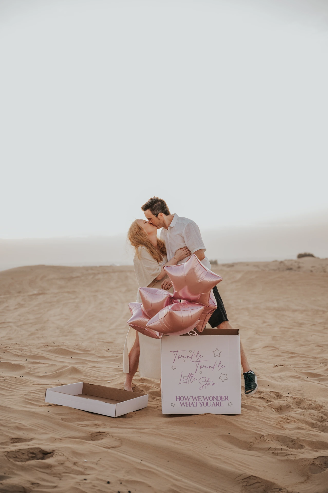 A couple stands together on Dubai desert dunes opening a box with pink star balloons during a gender reveal photoshoot.