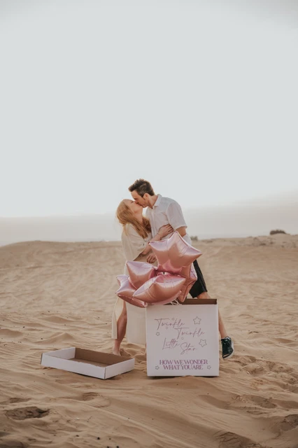 A couple stands together on Dubai desert dunes opening a box with pink star balloons during a gender reveal photoshoot.