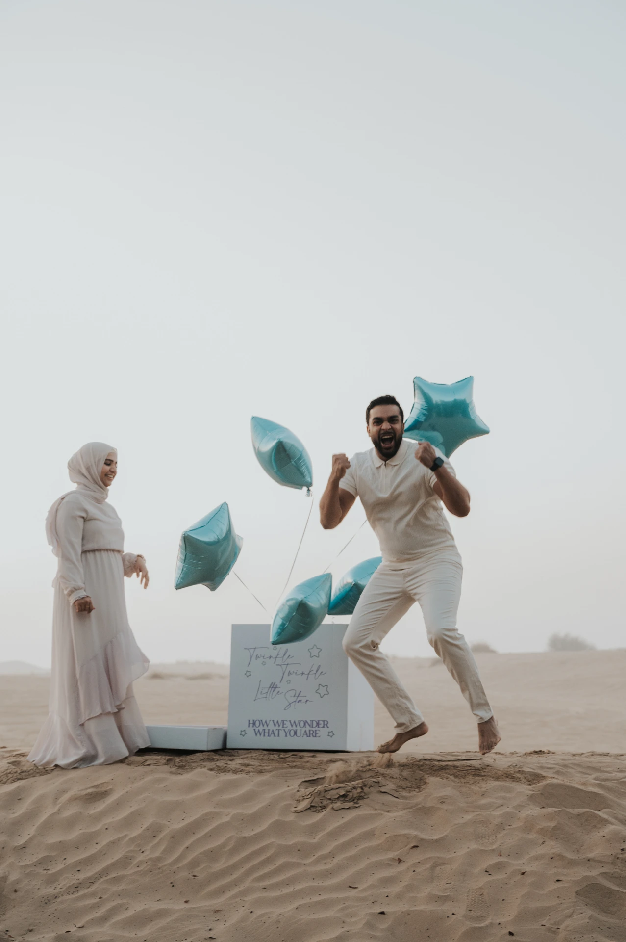A father laughs while holding blue star balloons as his partner watches on Dubai desert dunes during a playful gender reveal photoshoot.