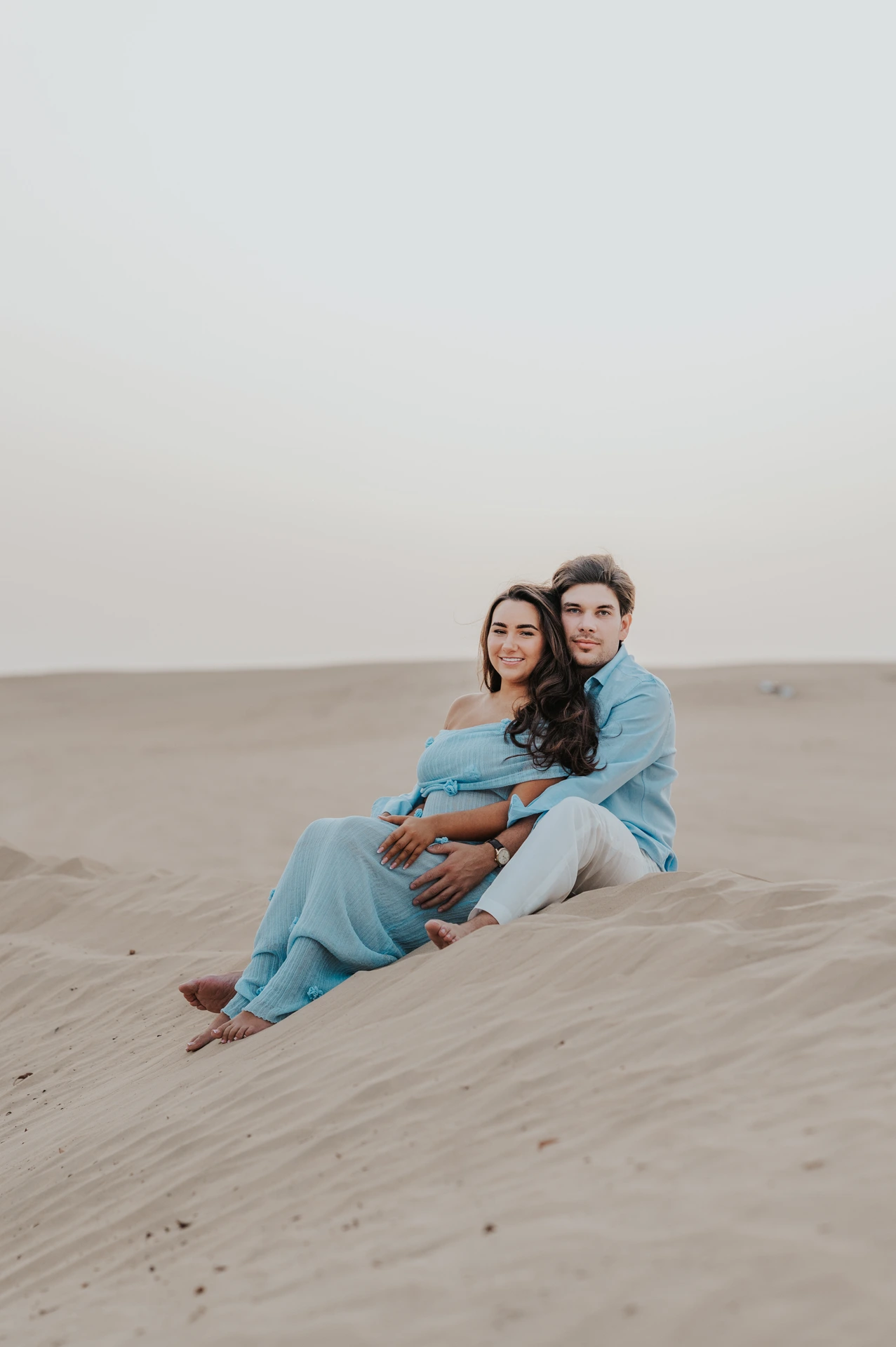 Gender Reveal Photoshoot Dubai - seated couple dunes portrait A couple in blue outfits sits close together on Dubai dunes during a soft and intimate gender reveal photoshoot portrait.