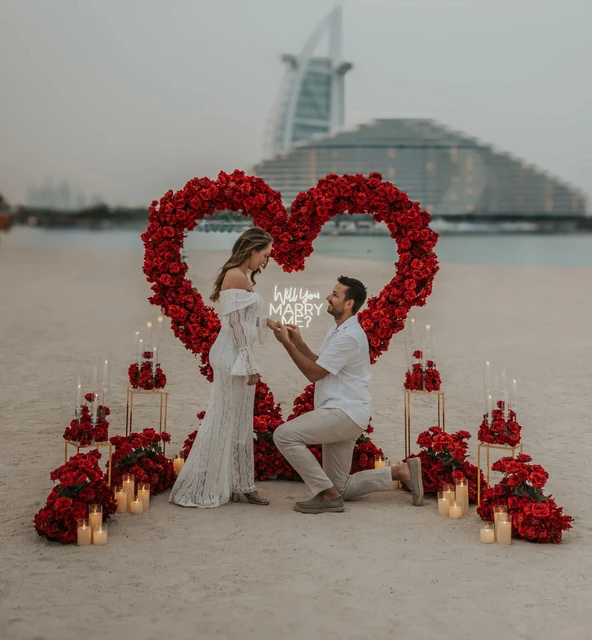 A man kneels to propose on a Dubai beach beside a red floral heart setup, with candles lit and Burj Al Arab visible behind the couple.