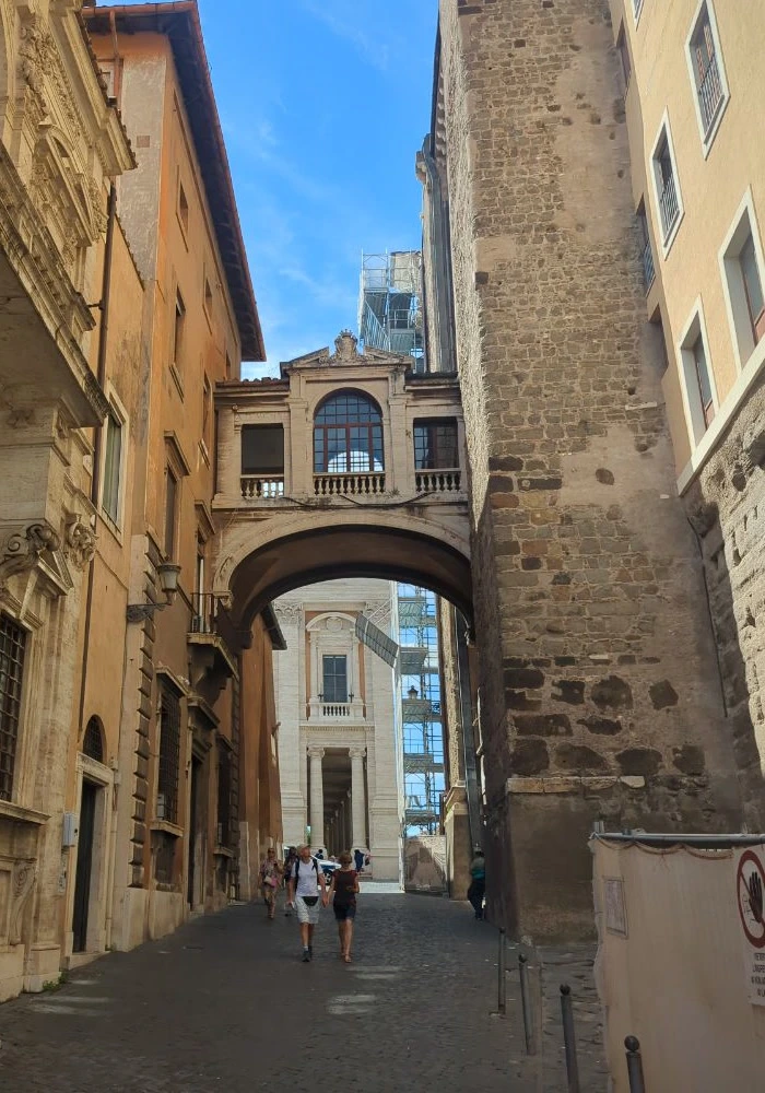 Visitors walk along a shaded cobbled lane beneath an overhead bridge, with warm stone facades framing a portrait setting in Rome.
