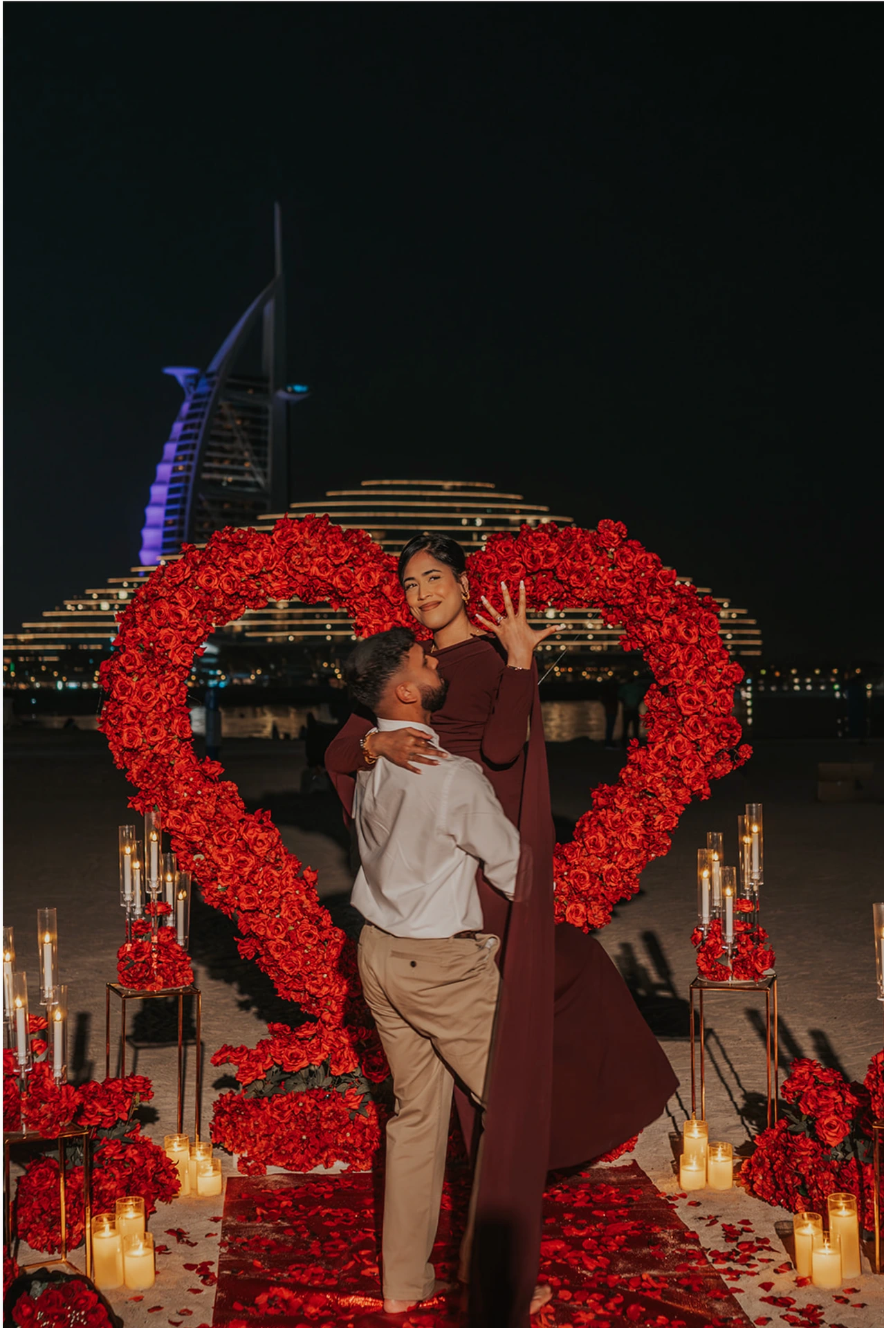 A couple embraces inside a red heart proposal setup on Dubai beach at night, with candles and Burj Al Arab lights behind them.