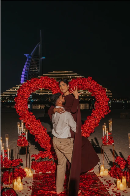 A couple embraces inside a red heart proposal setup on Dubai beach at night, with candles and Burj Al Arab lights behind them.